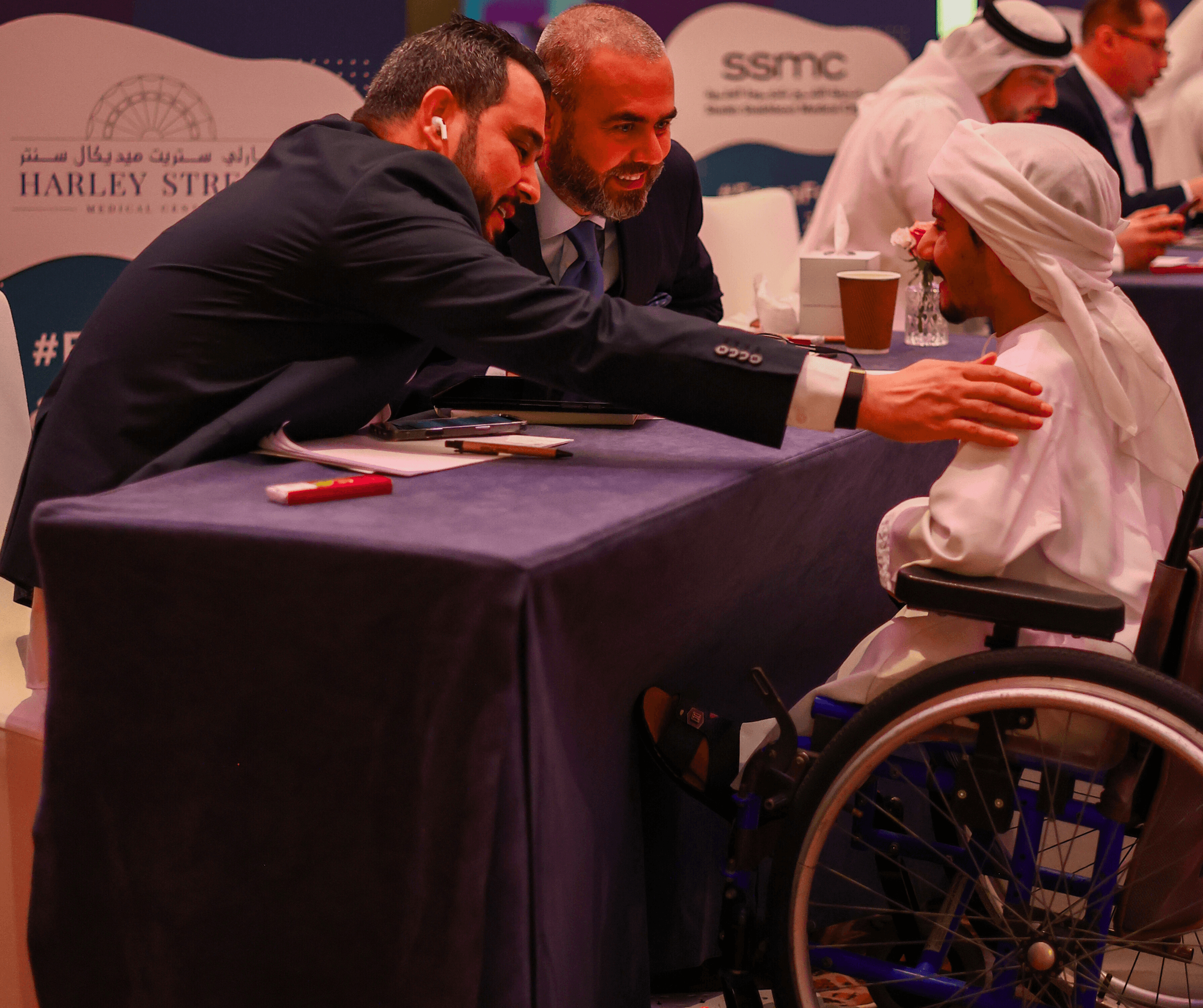 Two businessmen interacting with a young person in traditional dress using a wheelchair at an event.