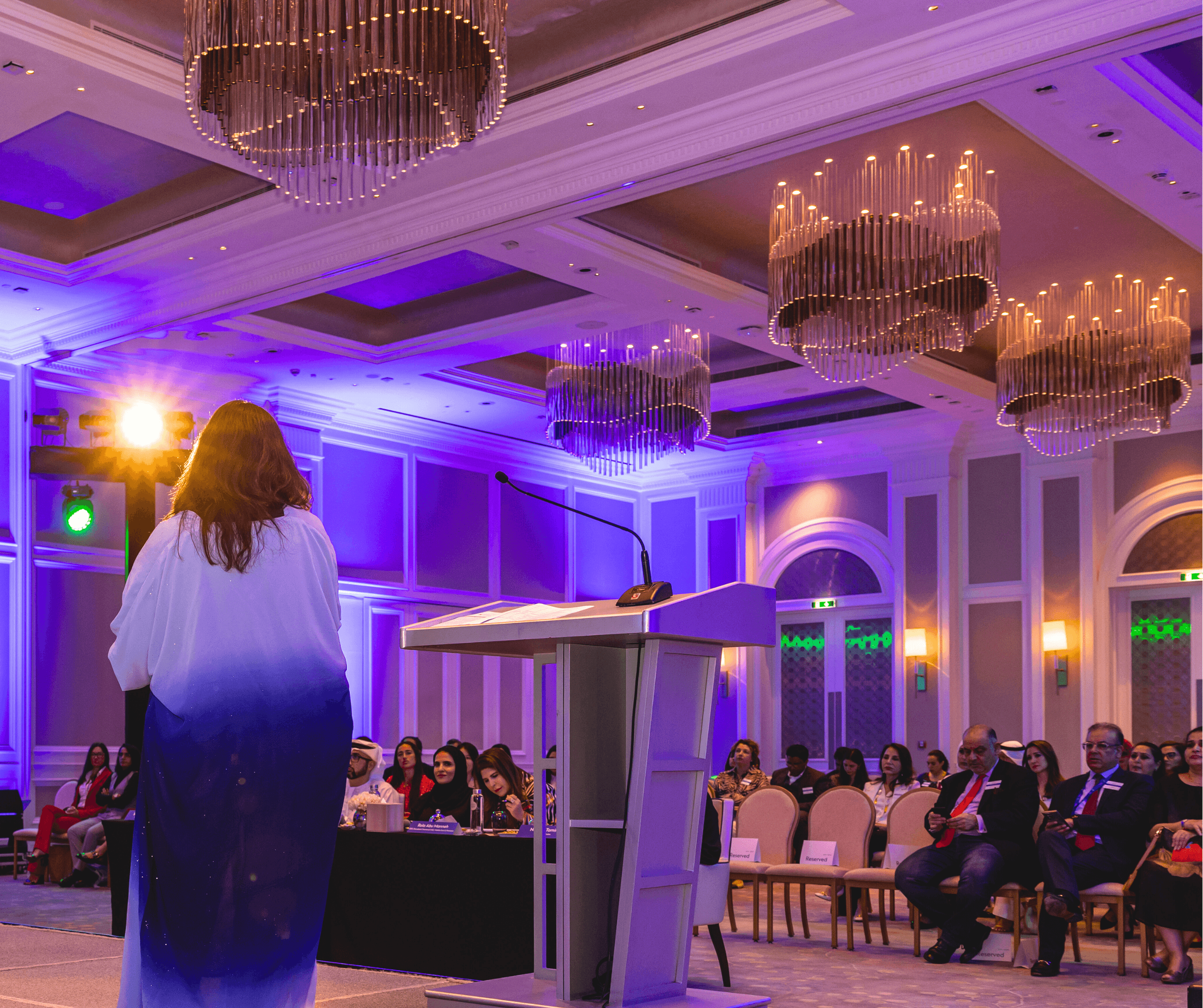 View from behind a presenter facing an audience in an elegant ballroom with purple lighting and chandeliers.