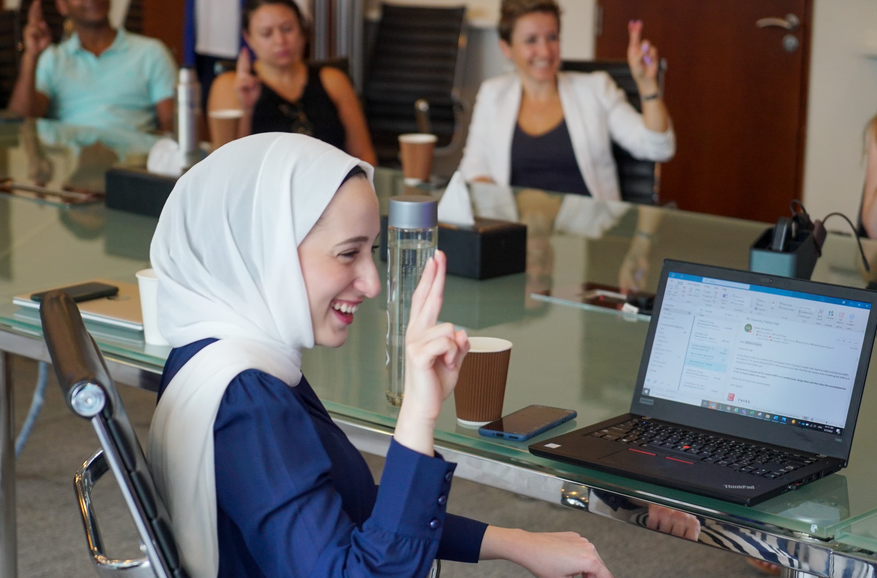 Woman in white hijab smiling during a video meeting at a glass desk with laptop and coffee cup.